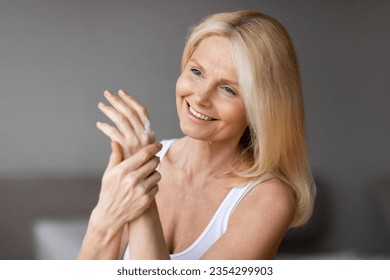 Skincare for hands. Happy middle aged woman applying moisturizing cream to her hands, enjoying her soft skin, sitting in bedroom interior and smiling, copy space - Powered by Shutterstock