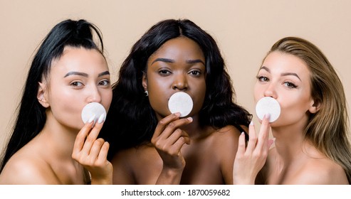 Skincare And Natural Beauty Concept. Three Diverse Women With Nude Makeup, Asian, African And Caucasian, Holding Cotton Pads, Covering Their Lips, Standing On Beige Background, Close Up
