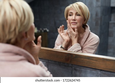 Skincare Routine. Mature Woman Touches Face Skin In Front Of The Mirror Reflection. Anti Aging Beauty Treatment Concept.