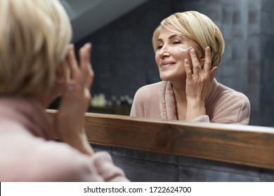 Skincare Routine. Mature Woman Uses Cosmetic Cream On Face Skin In Front Of The Mirror Reflection. Anti Aging Beauty Treatment Concept.