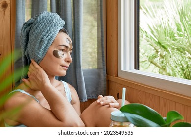 Skincare Routine. Middle Age Woman With Wrinkles And Hydrogel Under-eye Patches Drinking Smoothie And Looking In Window. Lifting Anti-wrinkle Mask Under Eyes. Skincare, Collagen Mask And Spa Concept.