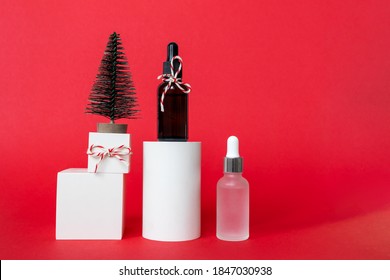 Skincare Serum And Essence In Glass Bottle With Pipette, Gift Box And Christmas Tree Decor On White Geometric Podium On Red Background. Facial Skincare Winter Holidays Modern Composition