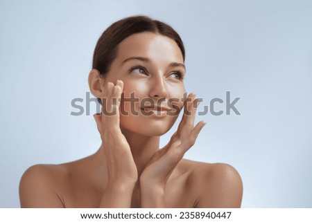 Skincare. Woman with beautiful face touching healthy facial skin. Beautiful portrait of smiling Asian girl model with natural makeup enjoys glowing hydrated skin on blue background closeup. High