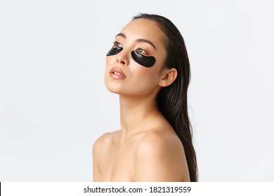 Skincare, women beauty, hygiene and personal care concept. Profile of attractive sensual asian woman standing naked and looking away, touching clean skin, using eye patches, remove dark circles