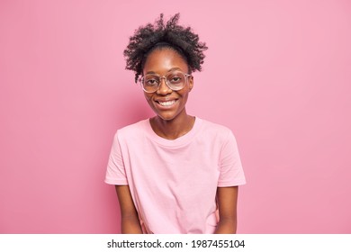 Skinny dark skinned woman with curly hair smiles broadly enjoys leisure free time wears big transparent glasses t shirt isolated over pink background. Peope ethnicity sincere emotions concept