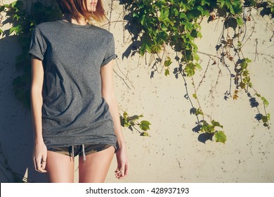 Skinny girl in a gray blank t-shirt is looking aside. A young model look woman is standing beside the wall overgrown with ivy.