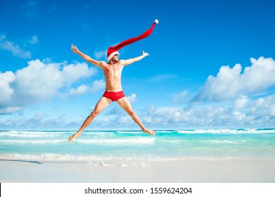 Skinny man with extra long Santa hat doing a Christmas celebration star jump in red swimming briefs on the shore of a tropical beach