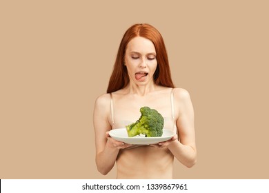 Skinny woman with ginger long hair, dressed only in underwear, looking at piece of fresh broccoli with adoration, licking, wants to eat it very much