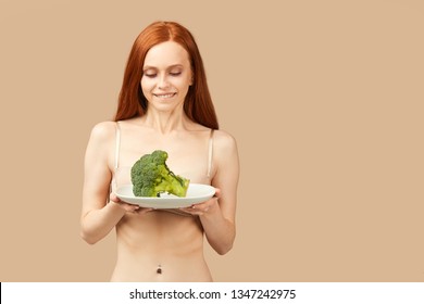 Skinny woman with ginger long hair, dressed only in underwear, looking at piece of fresh broccoli with adoration, licking, wants to eat it very much