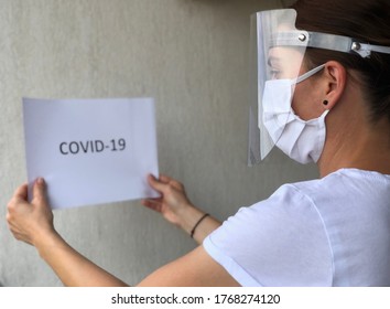 Skopje, Macedonia- July 2,2020: Woman Working Protected With White Medical Mask And Plexiglass Face Shield From Coronavirus Covid-19 And White Paper With Text Covid-19