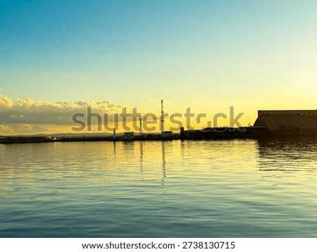 The sky changes color at sunset, casting light on the water. Structures stand by the shore, reflected in the still water of the port. Clouds drift along the horizon.