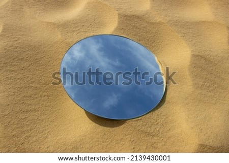 The sky with clouds is reflected in a round mirror, which lies on a sandy surface