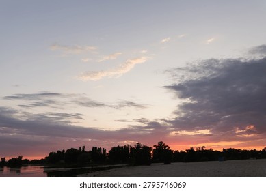 The sky displays a gradient of soft pinks and oranges as clouds drift across the horizon above a calm body of water and a dark treeline