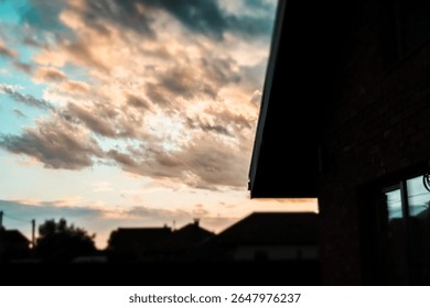 The sky with dramatic clouds bathed in warm glow of sunset, over houses. A dramatic sky of layered clouds glows in the warm light of sunset, framed by the silhouetted edge of a building and rooftops.