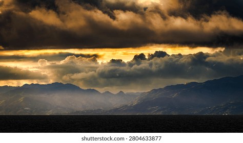 The sky is filled with dramatic, dark clouds illuminated by a golden sunset, casting a moody glow over the distant mountains and sea