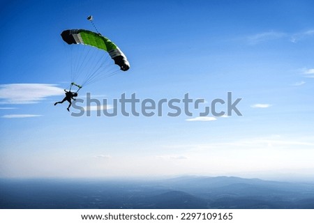 Skydiver flying a green parachute silhouetted against blue sky and clouds over rural fields and mountains