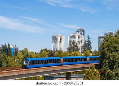 Skytrain moves along its elevated track in Vancouver, Canada. The train is surrounded by trees and tall buildings under a bright blue sky. It shows public transportation in action.