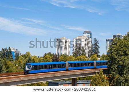 Skytrain moves along its elevated track in Vancouver, Canada. The train is surrounded by trees and tall buildings under a bright blue sky. It shows public transportation in action.