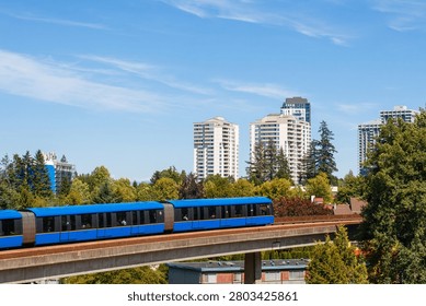 The Skytrain moves smoothly along its elevated track in Vancouver, Canada. Surrounding buildings stand tall under a clear blue sky. Trees and green spaces add to the urban scene.