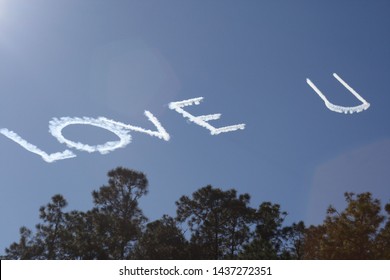 Skywriting Quote Love You. Plane Written Love U Against Blue Sky Visible Above Green Trees. Affectionate Writing Brings Happiness. Makes You Think Of Valentines Day, Anniversary, Etc. Sky Writing