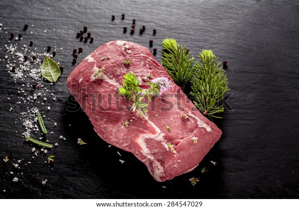 Slab of uncooked wild boar\
steak for roasting on a barbecue seasoned with spices and herbs\
lying alongside a small fir branch on a dark background, overhead\
view