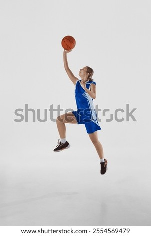 Slam dunk. Full-length vertical image of young teen girl in blue uniform, basketball player in motion, playing, training against white background. Concept of sport, youth, motion, healthy lifestyle