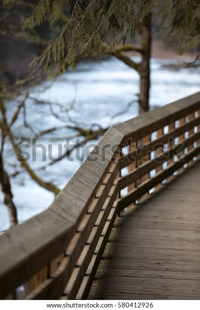 Slanted wood railing of plank river boardwalk\
path with trees and river in\
background