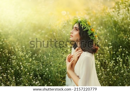 slavic fantasy young woman praying, hands folded in prayer pose. White traditional dress, herbal wreath on head. Nature summer, green grass, divine magic light, bright sun. Girl holiday Ivan Kupala