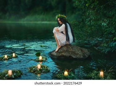 Slavic girl sits on stone on shore lake. Nymph fantasy woman hugs knees. Long black hair. Wreaths of grass, flowers float on water. Candles burning. River dusk forest green tree. Riutal of Divination. - Shutterstock ID 2022176429