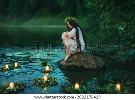 Slavic girl sits on stone on shore lake. Nymph fantasy woman hugs knees. Long black hair. Wreaths of grass, flowers float on water. Candles burning. River dusk forest green tree. Riutal of Divination.