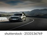 A sleek black sedan navigates a sharp curve on a mountain road, surrounded by rolling green hills under a cloudy sky