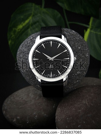 a sleek black wristwatch with a black dial and silver ring is showcased on a black leather belt, resting on a stone surface with a backdrop of green leaves.