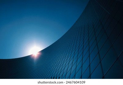 The sleek, curved facade of a modern office building in blue sky. The sun peeks from behind the structure, creating a captivating play of light and shadow on the building's reflective glass surface. - Powered by Shutterstock