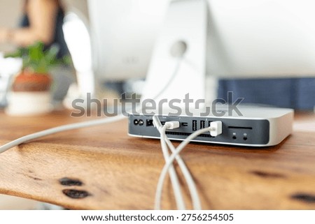 A sleek desktop computer setup on a wooden desk, featuring a monitor and a compact CPU with various cables connected. A plant is blurred in the background.