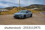 A sleek gray sedan parked on a gravel pull-off by a rural road, set against rolling hills and golden fields under an overcast sky. The scene captures freedom, adventure, and modern automotive design.