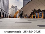 A sleek pedestrian walkway with orange metal arches curving overhead, leading towards tall urban buildings under a cloudy sky. A person walks away in the distance.