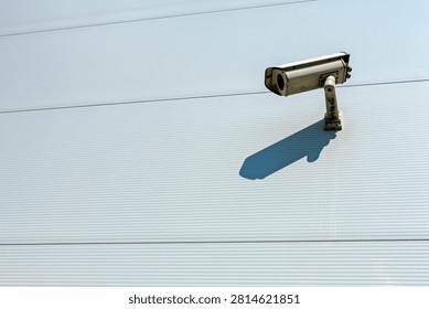 A sleek security camera is mounted on a corrugated light metal wall, casting a sharp shadow under bright direct sunlight. In the upper right quadrant of the composition, an outdoor closed-circuit television device extends horizontally from a sturdy mounting bracket fastened securely to an exterior wall surface. The background consists of uniform, horizontal cladding panels featuring subtle, finely spaced ribbed textures running end-to-end across the frame, interrupted only by thin parallel seams separating individual sections. Sunlight strikes the scene from an elevated angle above and behind the apparatus, producing an elongated silhouette angled downward across the metallic paneling. The camera housing exhibits a boxy, industrial profile encased in a weathered off-white protective shell designed to endure various atmospheric conditions. Tiny traces of dirt, grime, and minor discoloration along the joints and mounting base hint at prolonged exposure to environmental elements over time. Its circular glass lens aperture faces forward with an uncompromising view, serving as a vigilant ocular instrument for constant monitoring and protection. The striking contrast between the sun-drenched, pale grey cladding and the dark, precise outline of the cast shadow creates a strong visual dynamic characterized by geometric simplicity and stark minimalism. Every mechanical detail, from the rear cable connectors to the adjustable swivel joint, underscores the functional nature of commercial surveillance architecture. Light reflects smoothly off the top curved edge of the visor hood, highlighting its sleek contours against the subtle rhythmic pattern of the background structure. Overall, the scene conveys a calm yet watchful atmosphere, combining modern urban security infrastructure with clean lines, high contrast illumination, and quiet structural order.