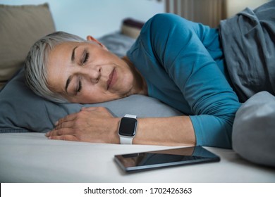 Sleep Apps - Tech-savvy Senior Woman Sleeping In Bed, Using Smart Phone And Smart Watch To Improve Her Sleeping Habits