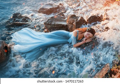 sleep beauty charming sea princess resting on wet stones, girl in blue long tender dress lies in cold water waterfall hands under head red hair, young woman river nymph mermaid cute face. rays sun 