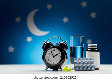 Sleep Disorder, Bedtime And Medicine Concept - Close Up Of Alarm Clock, Glass Of Water, Earplugs And Sleeping Pills Over Moon And Night Stars On Blue Background