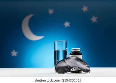 sleep disorder, bedtime and object concept - close up of eye mask, glass of water and soporific medicine over moon and night stars on blue background