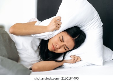 Sleep And Rest Concept. Sad Chinese Girl, Lying In A Bed At Home, Covers Her Ears With A Pillow, Tries To Sleep, Closes Herself From Noise, Has A Bad Dream, Restless Dream