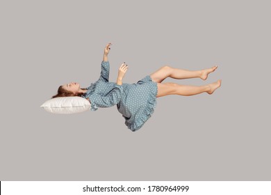 Sleeping Beauty Floating In Air. Relaxed Girl In Vintage Ruffle Dress Keeping Eye Closed, Lying On Pillow Levitating, Flying In Dream With Hands Up To Catch. Studio Shot Isolated On Gray Background