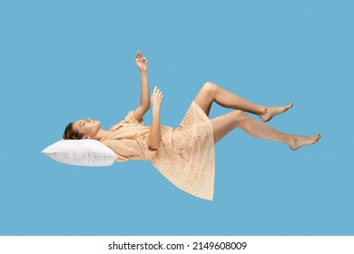 Sleeping beauty floating in air. Relaxed girl in yellow dress keeping eye closed, lying on pillow levitating, flying in dream with hands up to catch. studio shot isolated on blue background
