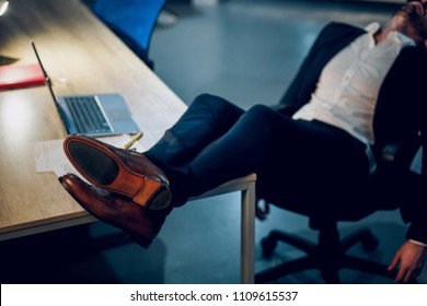 Sleeping businessmans feet up on office table. Cropped picture of young mans feet with brown leather shoes on lying on office desk with laptop in background.