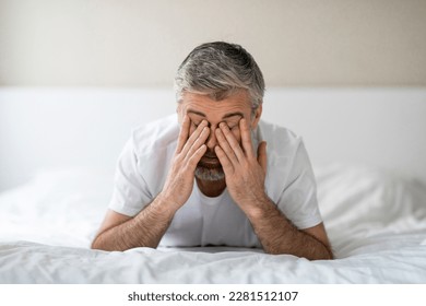 Sleeping disorder. Closeup of exhausted grey-haired middle aged man lying in bed in the morning, rubbing eyes, feeling tired after sleepless night, white bedroom interior, copy space