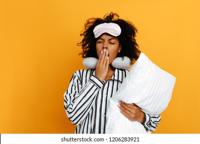 Sleeping. Dreams. Woman Portrait. Afro American Girl In Pajama And Sleep Mask Is Holding A Pillow And Yawning, On A Yellow Background