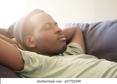 Sleeping handsome black man with full lips wearing black hat and green T-shirt having good dreames being tired after work. Dark-skinned hipster sleeping at cozy sofa at home. People and rest