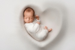 A Sleeping Newborn Boy In The First Days Of Life In A White Soft Cocoon With A Knitted Woolen White Hat On A White Background In The Shape Of A Heart. Studio Macro Photography, Portrait Of A Newborn. 