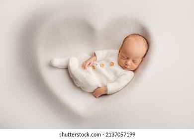 A sleeping newborn boy in the first days of life in a white soft cocoon with a knitted woolen white hat on a white background in the shape of a heart. Studio macro photography, portrait of a newborn. - Shutterstock ID 2137057199